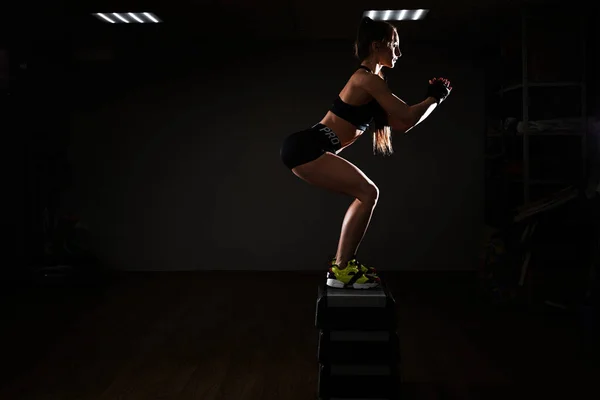 Fit young woman doing a step jump exercise. Muscular woman doing box squat at the gym on dark backround