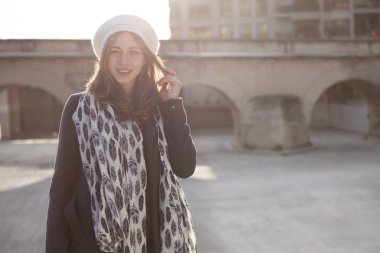 Kışlık kıyafetler giyen genç bir kadın gün batımında arka planda bir Roma köprüsü ile şehirde yürüyor.