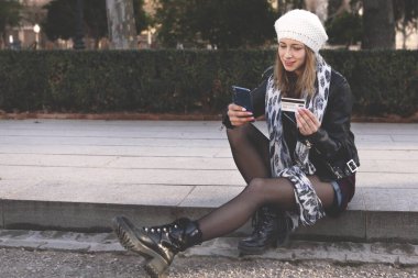 Granada 'da arka planda parkı olan bir sokakta oturan genç ve güzel bir kadın kredi kartı ve telefonuyla internetten alışveriş yapıyor.