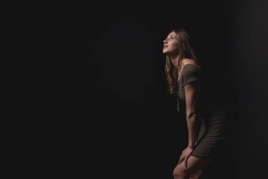 Siyah bir arka planda duvara yaslanmış güzel, genç bir model yukarıdan gelen ışıkla, gündelik ama zarif bir tarzla.