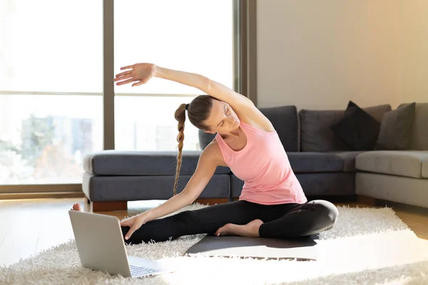 Çevrimiçi spor fitness yoga eğitimi. Yoga minderi üzerinde laptopla egzersiz yapan bir kadın. Evde online eğitim görüyor.