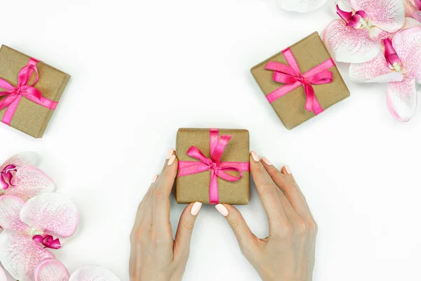 Flat-lay de presente caixas com mãos femininas e fita rosa com flores de orquídea vista superior de fundo branco. primavera — Fotografia de Stock