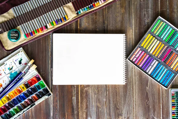 Düz sanat işyeri, tahta masa kalemler, fırçalar, suluboya boyalar, kağıt ve pastel renkler üst görünüm.