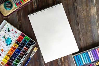 Sanat eserleri, kalemler, fırçalar, suluboya boyalar, kağıt ve pastel tebeşirler. Tahta masanın üzerinde yatıyordu..