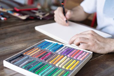 Tahta masadaki beyaz bir kağıt üzerinde pastel boya kalemi ile resim yapan kişi, sanatçı.