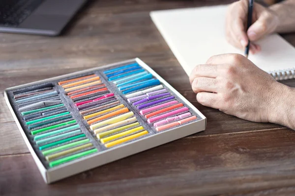 person painting with a pastel crayon chalk on a white sheet of paper with pallette of pastel chalks on the wooden table.