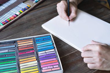 Beyaz bir kağıt üzerinde pastel boya ve boya kalemleriyle resim yapan bir adam. Tahta masadaki pastel tebeşir ve renkli kalemler.