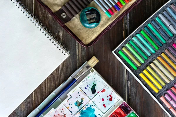 Arte lugar de trabajo, lápices de color, pinceles, pinturas de acuarela, papel y lápices de colores pastel. Colocación plana sobre mesa de madera — Foto de Stock