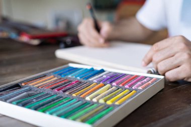 Beyaz bir kağıt üzerinde pastel renkli tebeşirle resim yapan kişi ve sanatçı önünde beyaz tebeşir lekesini ahşap masaya yapıştırıyor.
