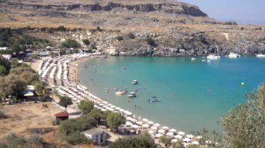 Lindos beach, Bay with clear water, sun umbrellas, people swimming, Mediterranean sea, yachts, rocky shore, Rhodes island Greece.