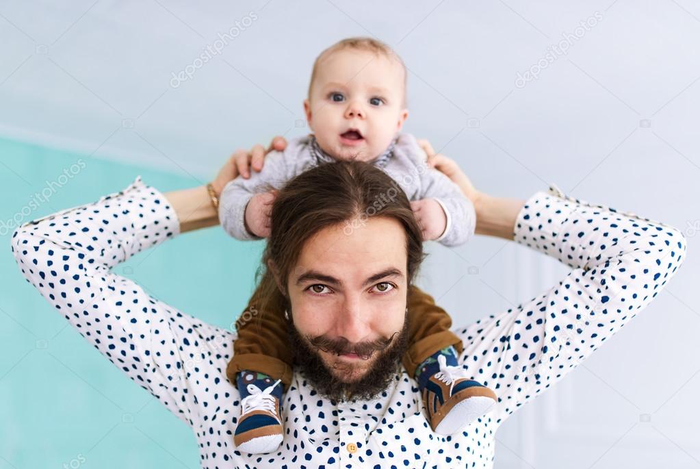 Charming baby son sitting on father neck in white room