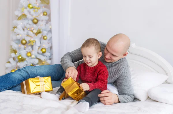 Padre e hijo pequeño yacen en la cama. Desempaquetan regalos de Navidad —  Fotos de Stock