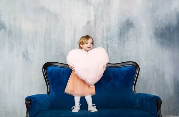 El encantador puesto de bebé en el sofá y tiene gran almohada de corazón en la mano . — Foto de Stock