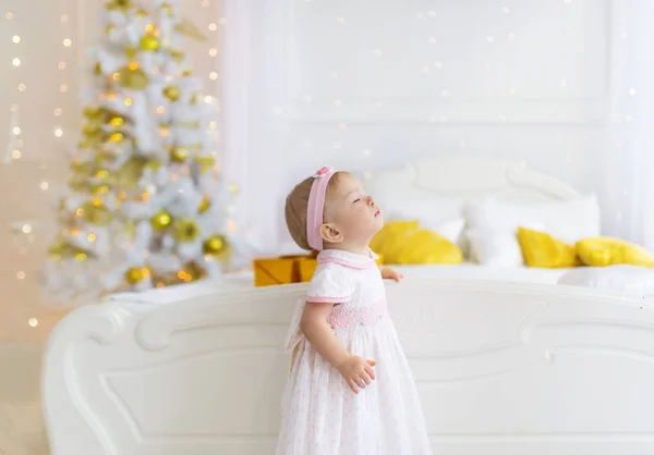 Adorable niña en un vestido blanco de pie en un piso cerca del árbol de Navidad y pide un deseo . — Foto de Stock