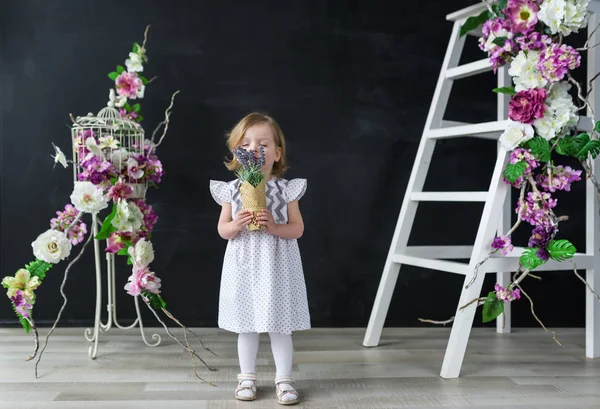 Adorável bebê menina vestindo um vestido branco cheirando belas flores — Fotografia de Stock