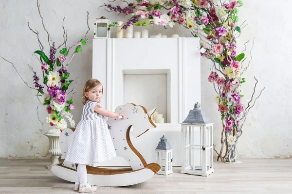 Retrato de uma menina loira em flores. Ela está perto do balancim cavalo de madeira na sala de flores brancas — Fotografia de Stock