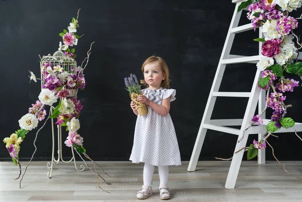 Petite fille en robe blanche. Bébé fille avec de belles fleurs debout dans la chambre des fleurs . — Photo