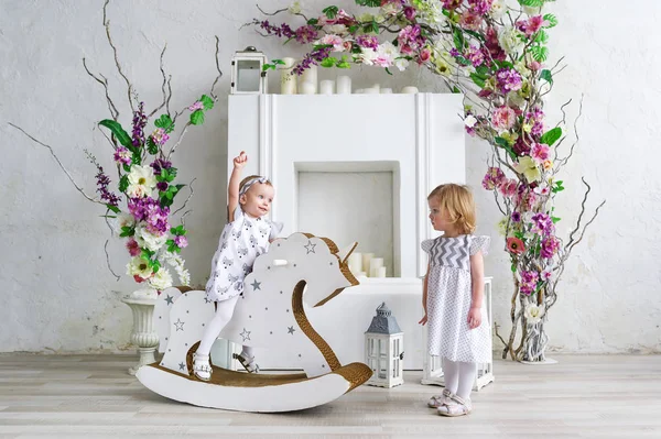 Deux charmantes petites filles jouent dans la salle lumineuse décorée de fleurs. Bébé fille balançant sur un cheval en bois — Photo