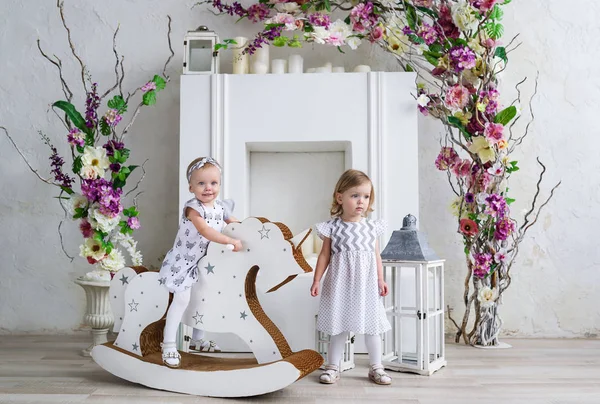 Zwei charmante kleine Mädchen spielen im hellen Raum, der mit Blumen dekoriert ist. Mädchen schaukelt auf einem Holzpferd — Stockfoto