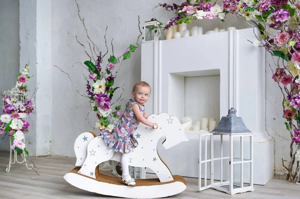 Linda niña montando un caballo de madera en la sala de flores — Foto de Stock