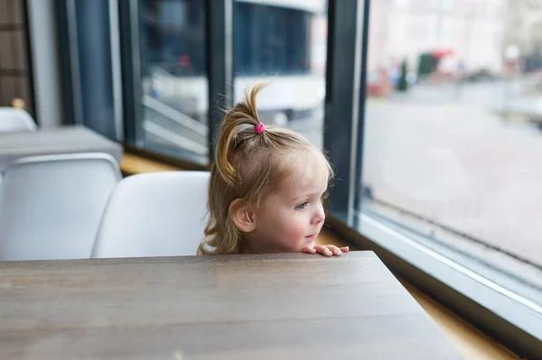 Маленька дівчинка дивиться з вікна, сидячи за столом у кафе — стокове фото