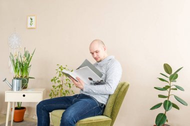 Kel adam koltukta otururken büyük kitap okuma portresi. Genç adam evde rahatlatıcı.