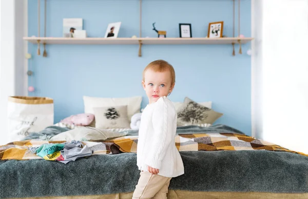 Cute pretty infant girl in a white sweater standing at a bed with pillows in bedroom. — Stock Photo, Image