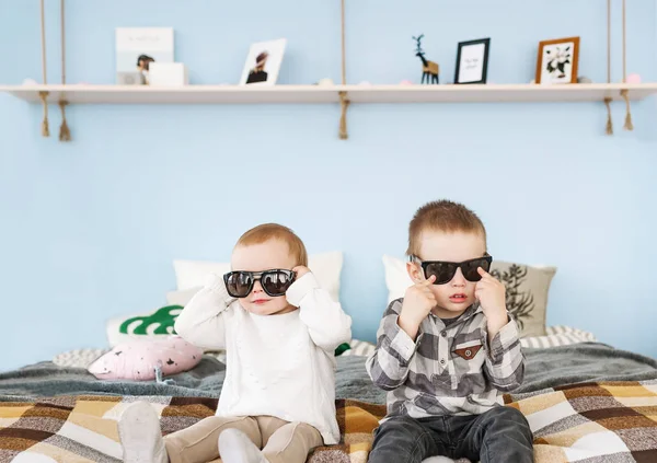 Portrait of brother and sister in sunglasses sitting on bed at home — Stock Photo, Image