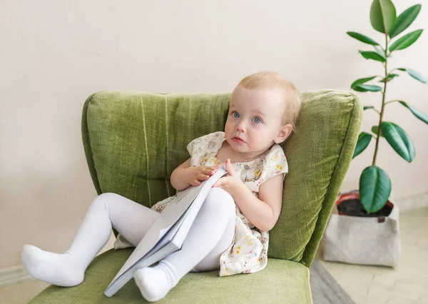 Linda niña pequeña hacer cara divertida, sosteniendo libro en la mano. Niño sentado en un sillón . — Foto de Stock