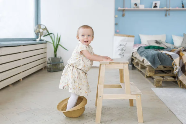 The cute baby girl stand on a straw hat and leans on a wooden chair — Stock Photo, Image