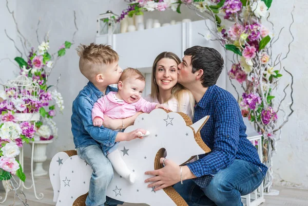 Familia feliz de cuatro sentarse en la habitación con flores y jugar con los niños en su caballo de juguete de madera — Foto de Stock