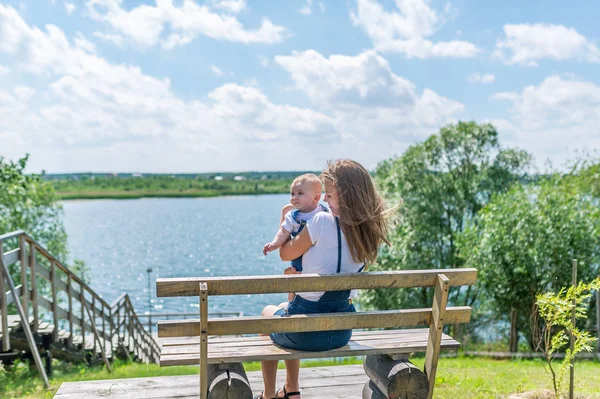 Young mother with infant sitting on the bench near a river — Stock Photo, Image