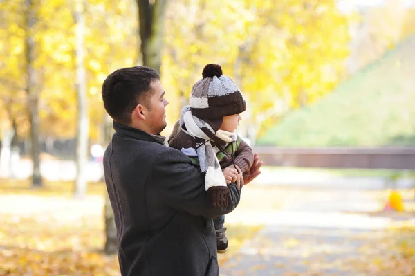 Счастливый папа держит своего сына, маленького смешного мальчика на руках, в холодный день, стоя вместе в осеннем парке . — стоковое фото