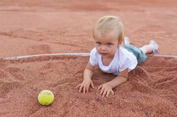 Маленька дівчинка лежить на тенісному корті. Маленька дівчинка і тенісний м'яч — стокове фото