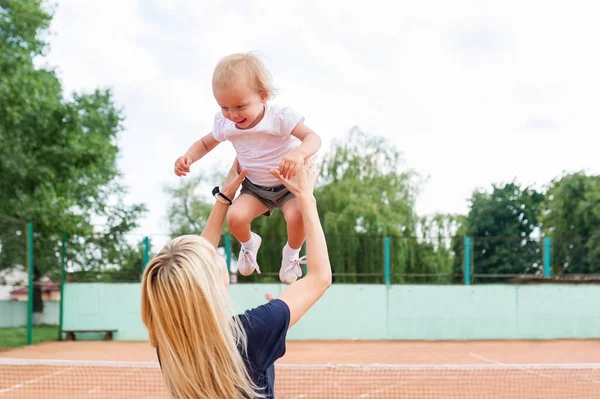 Hübsche Mutter lacht und zieht ihr kleines Mädchen mit Tennis im Hintergrund groß — Stockfoto