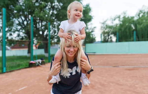 Petit enfant, une petite fille s'assoit sur les épaules de maman souriant, riant, jouant, s'amusant au court de tennis . — Photo