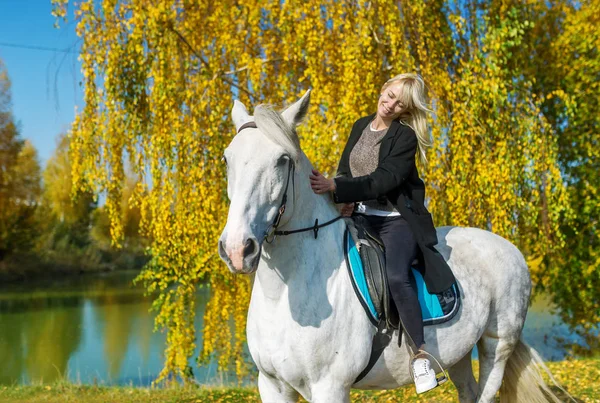 Ung kvinna rider en häst på en solig höstdag mot bakgrund av den blå sjön — Stockfoto