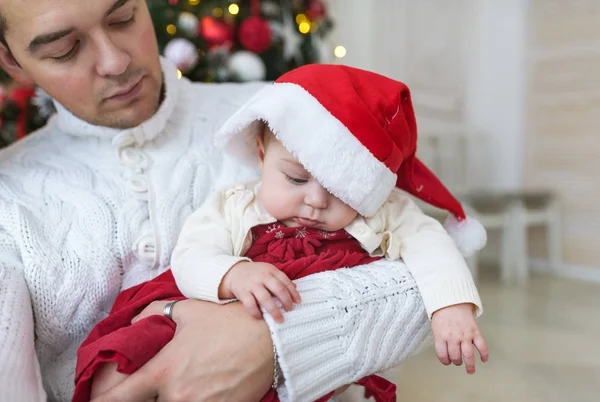 Retrato de primer plano del joven padre con ropa blanca sosteniendo a su bebé recién nacido contra un árbol de Navidad brillante decorado . — Foto de Stock