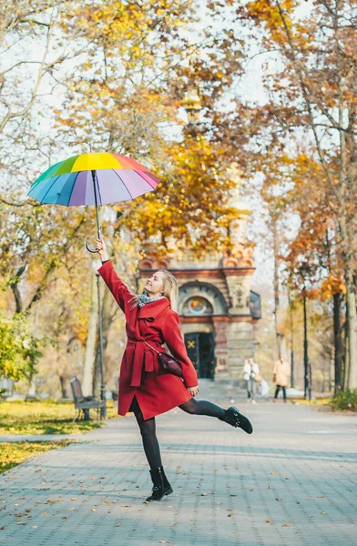 Jovem mulher de casaco vermelho com um guarda-chuva colorido arco-íris saltando ao longo da passarela de outono no parque da cidade — Fotografia de Stock