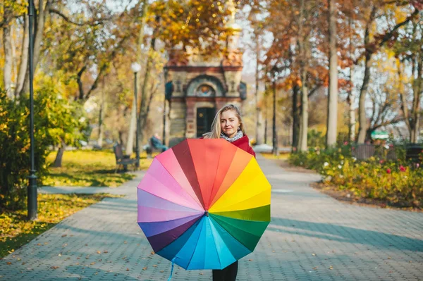 Un retrato de una hermosa joven en un parque de otoño de la ciudad con un paraguas brillante y colorido . — Foto de Stock