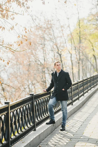 Un hombre con la mano en el bolsillo en un abrigo negro posa en el parque de otoño de la ciudad . —  Fotos de Stock