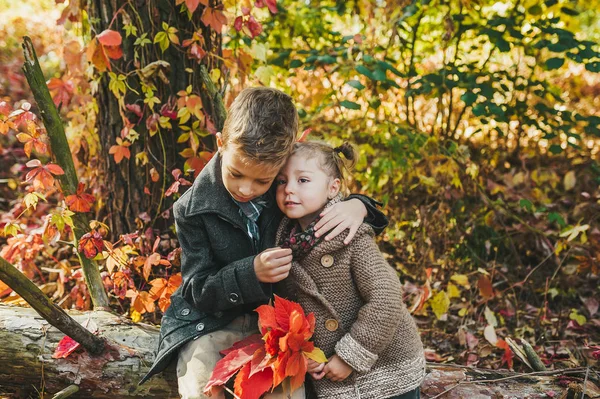 Niño y niña con hojas rojas en las manos sentados y abrazándose juntos en el tronco en el parque de otoño — Foto de Stock