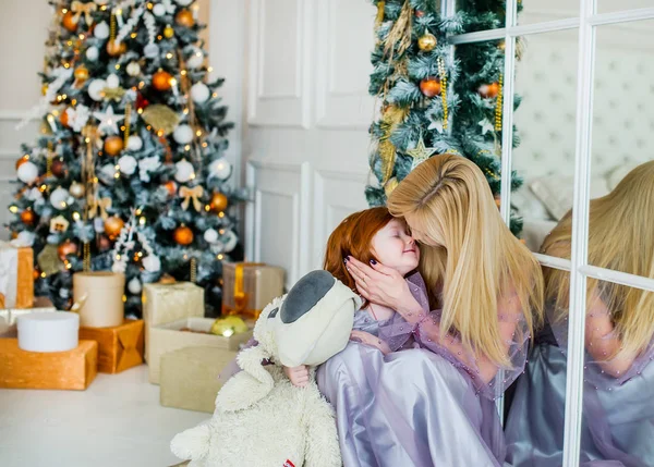 Mãe beijo ruiva pequena filha perto da árvore de Natal em casa . — Fotografia de Stock