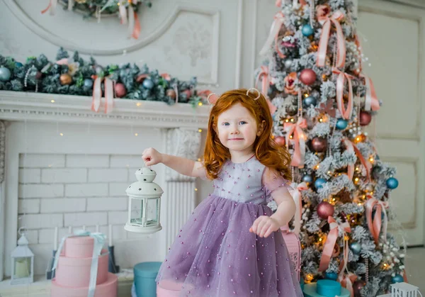 Pelirroja niña en un vestido violeta contra el telón de fondo de un árbol de Navidad con un candelabro blanco en la mano — Foto de Stock