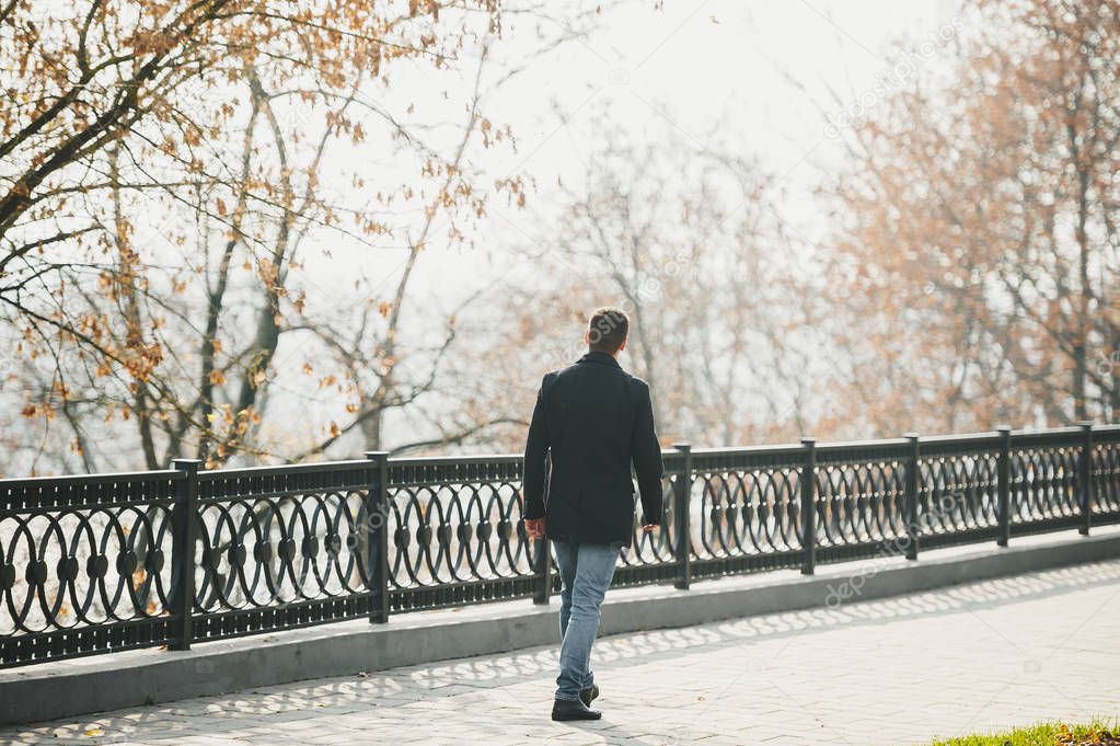 Handsome man in black coat walking around autumn city park.