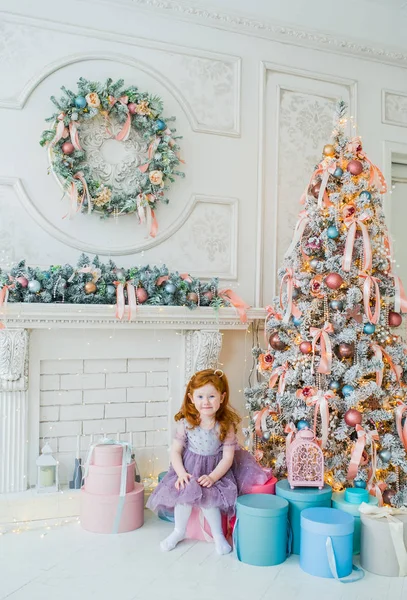 La niña pelirroja se sienta en una caja grande con regalos cerca del árbol de Navidad . — Foto de Stock