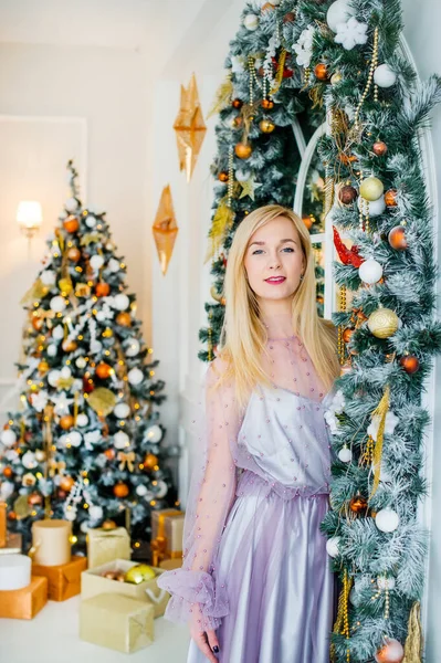 Блондинка в вечернем фиолетовом платье стоит у елки с подарками. Новый год — стоковое фото