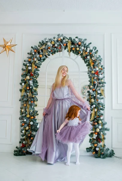 Woman with her little daughter in violet dresses dance before the fir arch decorated with Christmas accessories — Stock Photo, Image