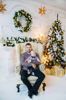 Genç baba koltukta oturuyor ve oğlunu yeni yıl ağacının yanında tutuyor.