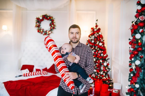 Papá sostiene al niño con un palo de juguete de pie ante un árbol de Navidad en el interior — Foto de Stock